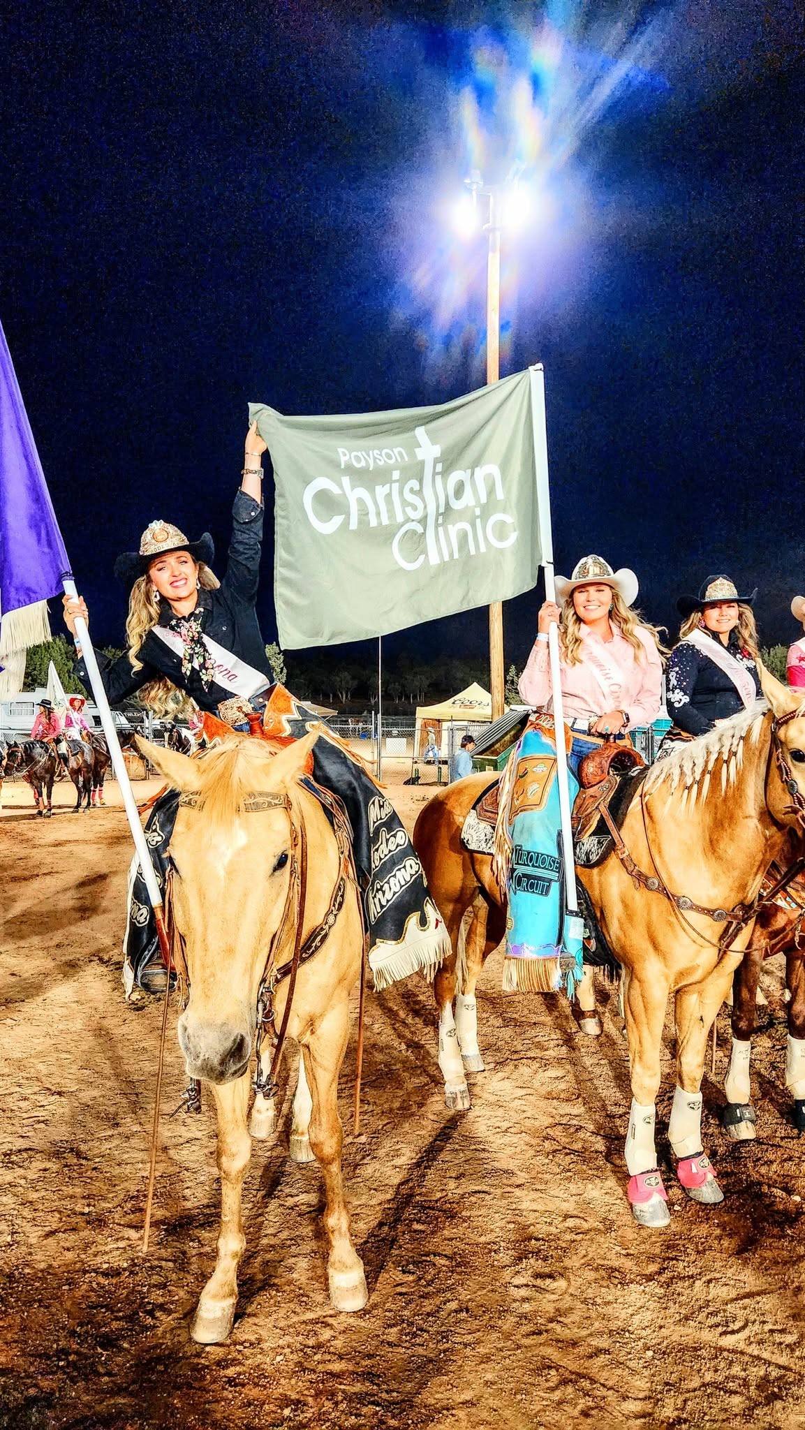 Rodeo royalty on horseback carrying a Payson Christian Clinic flag at night